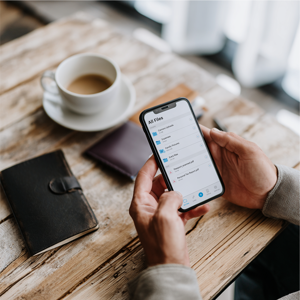 Hands holding a smartphone with Tresorit app open, checking secure files; coffee cup on table. Symbolizing safe and private digital life with Tresorit Secure Cloud for individuals.